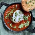 Shakshuka - placed in a cast iron skillet filled with jar sauce, a poached egg, and some greens with a slice of toasted bread topped with garlic oil and chopped basil