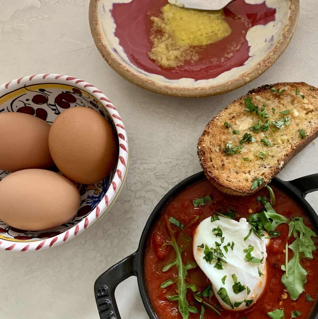 Shakshuka - placed in a cast iron skillet filled with jar sauce, a poached egg, and some greens with a slice of toasted bread topped with garlic oil and chopped basil. Also pictured 3 raw brown eggs in a bowl and a dish with fresh garlic oil