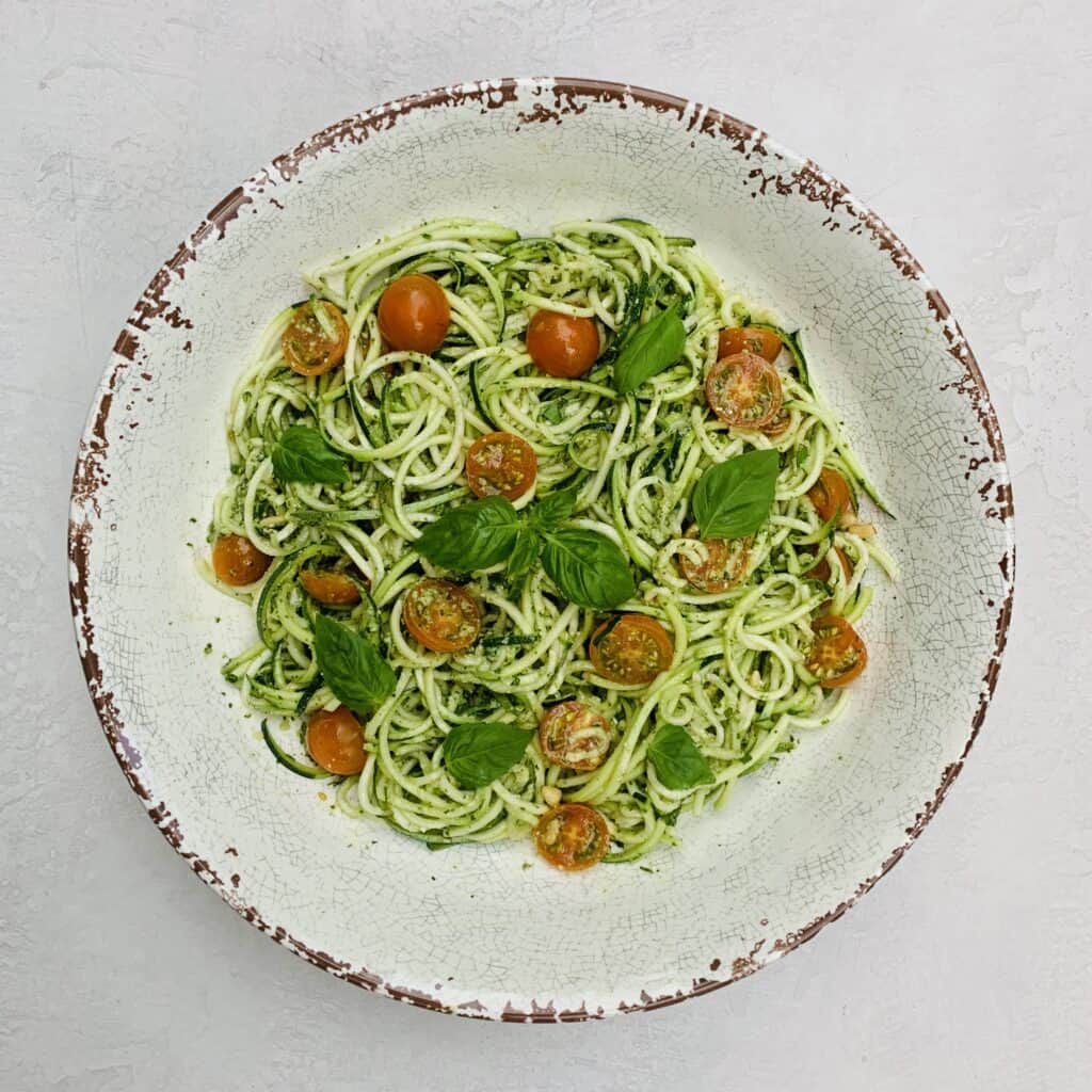 Pesto Zoodles and Tomato Salad pictured in a large serving bowl with pesto sauce in the middle and fresh basil leaves