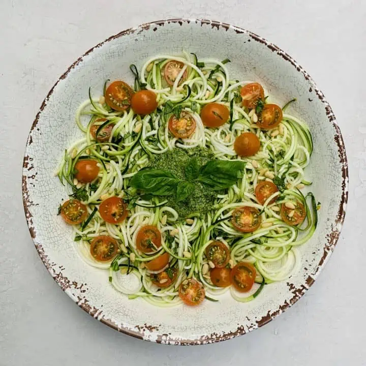 Pesto Zoodles and Tomato Salad pictured in a large serving bowl with pesto sauce in the middle and fresh basil leaves