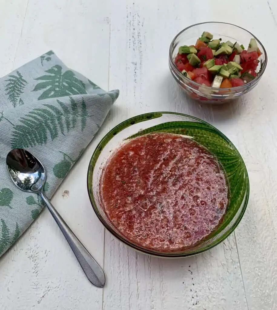 Bowl of Watermelon Gazpacho with toppings on the side of cucumber, avocado, red pepper, watermelon and herbs.