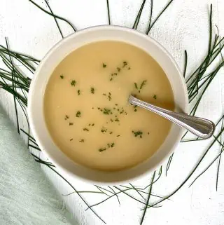 Vichyssoise Soup made with Potatoes, Cucumbers & Chives pictured in a bowl with scattered chives and a green napkin placed on a white wood board