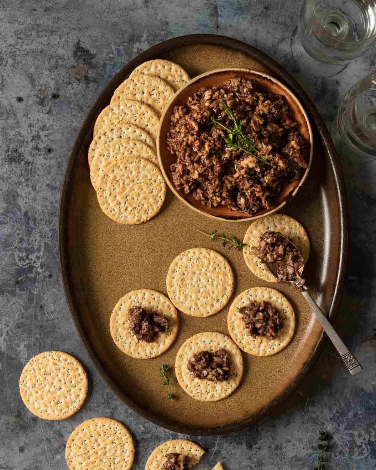 Brown dish of crackers being topped with mushroom tapenade. 