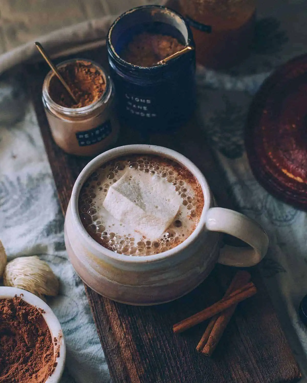 Mug of hot chocolate with a marshmallow next to jars of mushroom powder.
