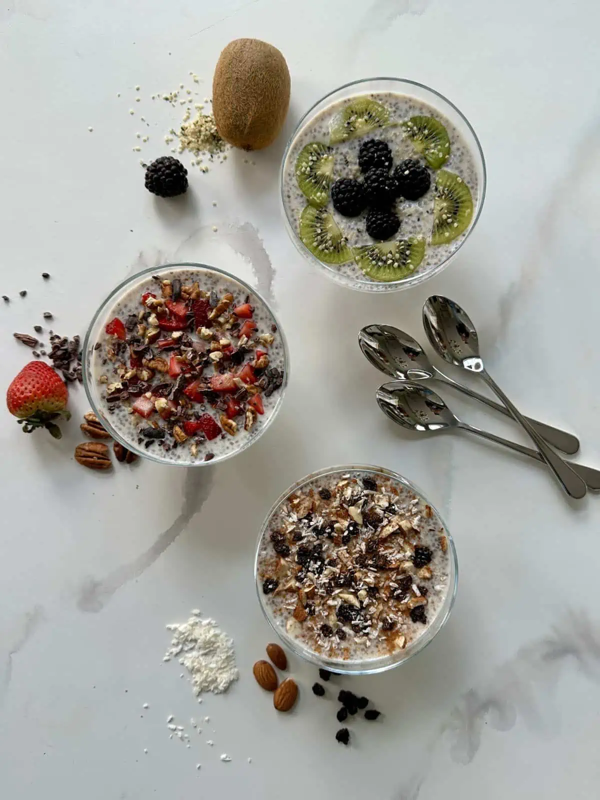 Three glasses of chia pudding with different toppings on the countertop with spoons next to them.