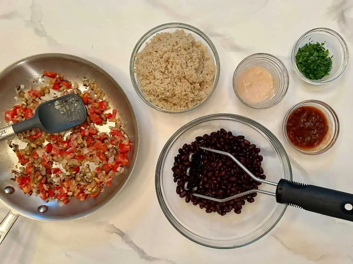 Black beans with a masher in a bowl surrounded by other ingredients that go in the patties.