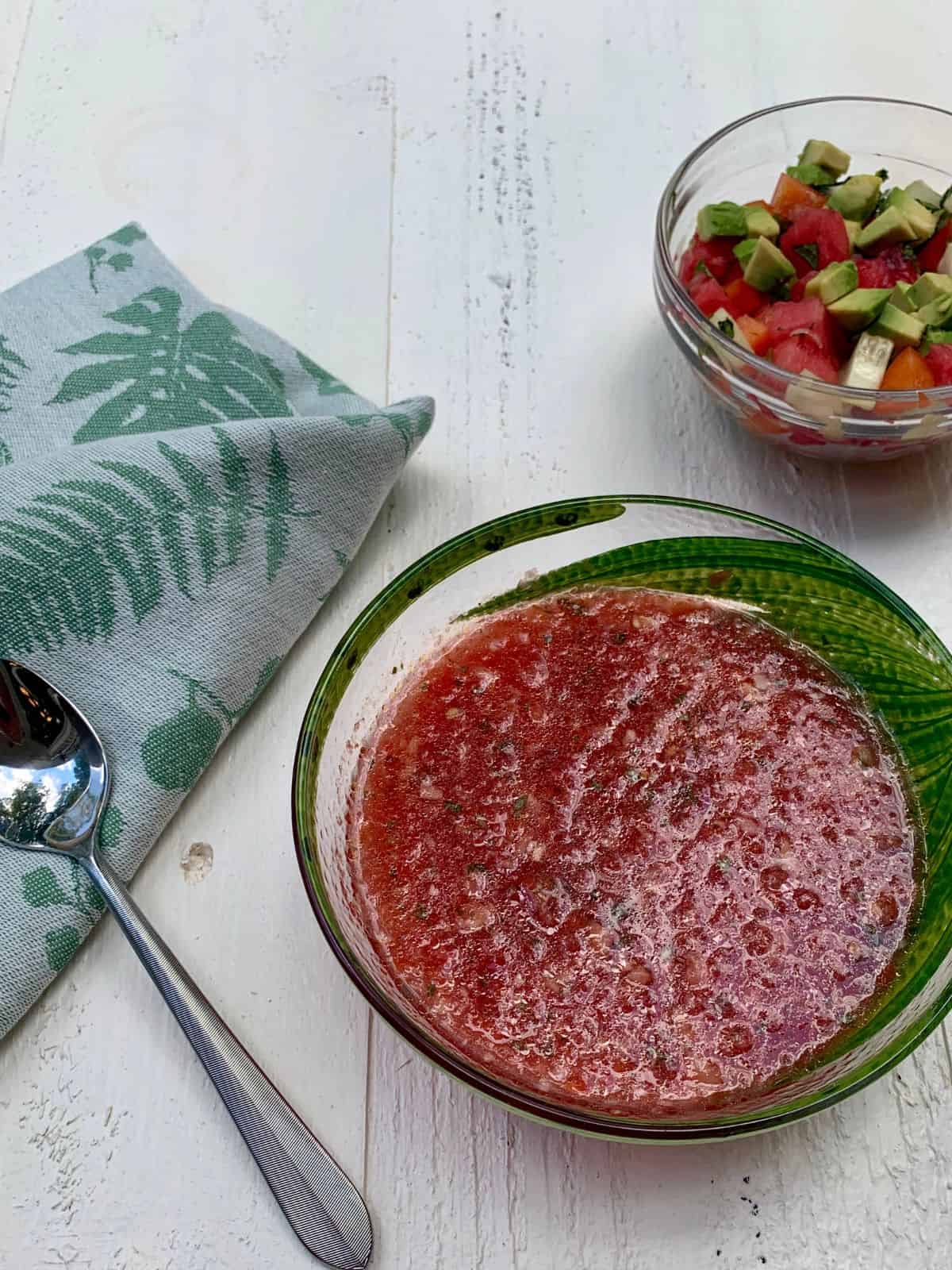 A bowl of watermelon gazpacho with a spoon on a napkin next to it and a bowl of toppings above the plate.