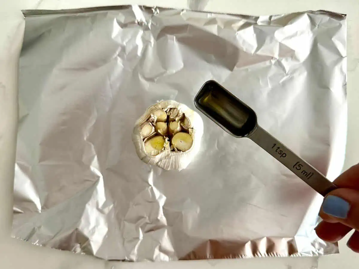 Head of garlic on foil with a hand holding a tablespoon drizzling with oil.